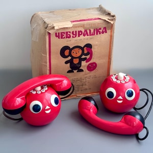 May include: A vintage red toy telephone with a round base and a handset, featuring large blue eyes and a smiling face. The box behind the phone has the text "Cheburashka" and a cartoon illustration.