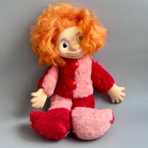 May include: A vintage doll with a painted face, orange curly hair, and a two-tone red and pink outfit. The doll's outfit includes a jacket with red buttons and matching pants and shoes. The doll is sitting on a gray surface.