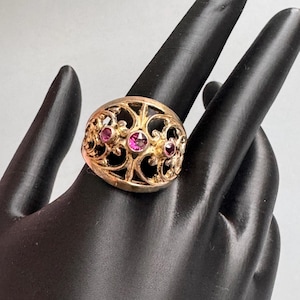 May include: A gold-toned ring featuring an intricate, openwork filigree design with scrolling metal patterns. Three small, round, vibrant pink gemstones are set horizontally across the center of the band, creating a focal point within the metalwork.