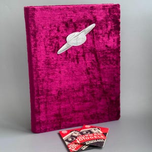 Puede incluir: Un álbum de fotos de terciopelo magenta con un emblema plateado que representa un globo terráqueo y alas. Dos pequeñas tarjetas rojas y blancas con texto e ilustraciones están en la parte inferior. El álbum es rectangular y parece vintage.