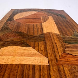 Peut inclure: Un plateau d&eacute;coratif carr&eacute; en bois avec un motif g&eacute;om&eacute;trique. Le plateau est compos&eacute; de diff&eacute;rentes teintes de bois, notamment marron clair, marron moyen et marron fonc&eacute;. Le motif pr&eacute;sente un cercle central et des formes angulaires.