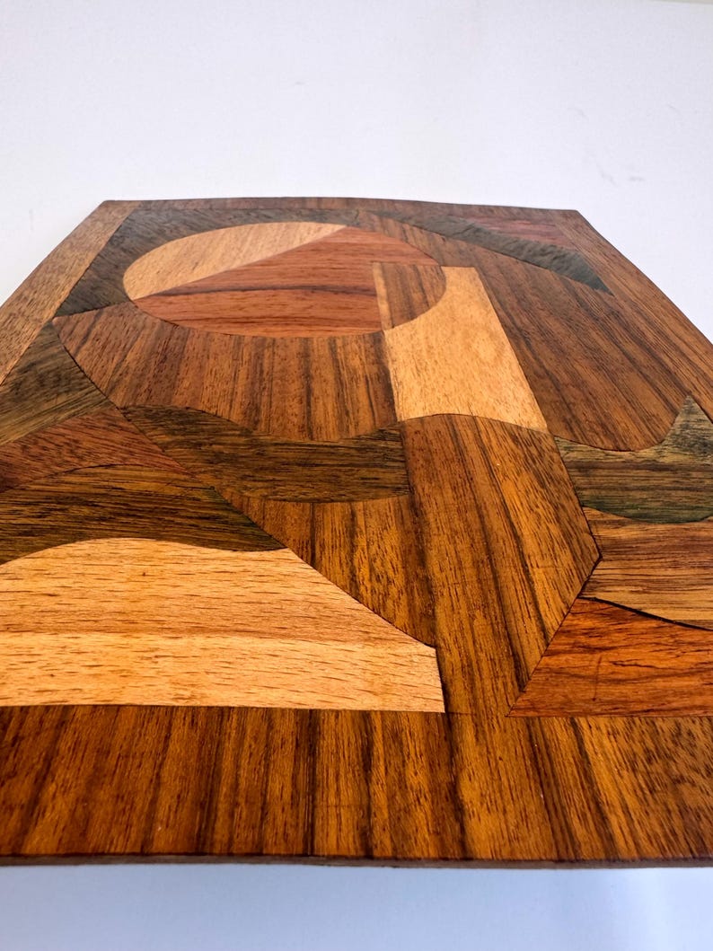 Peut inclure: Un plateau d&eacute;coratif carr&eacute; en bois avec un motif g&eacute;om&eacute;trique. Le plateau est compos&eacute; de diff&eacute;rentes teintes de bois, notamment marron clair, marron moyen et marron fonc&eacute;. Le motif pr&eacute;sente un cercle central et des formes angulaires.