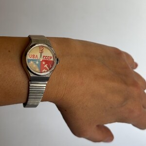 May include: A silver wristwatch with a white face and a design of the American and Soviet flags. The watch face reads "USA" and "CCCP" with a star and hammer and sickle.