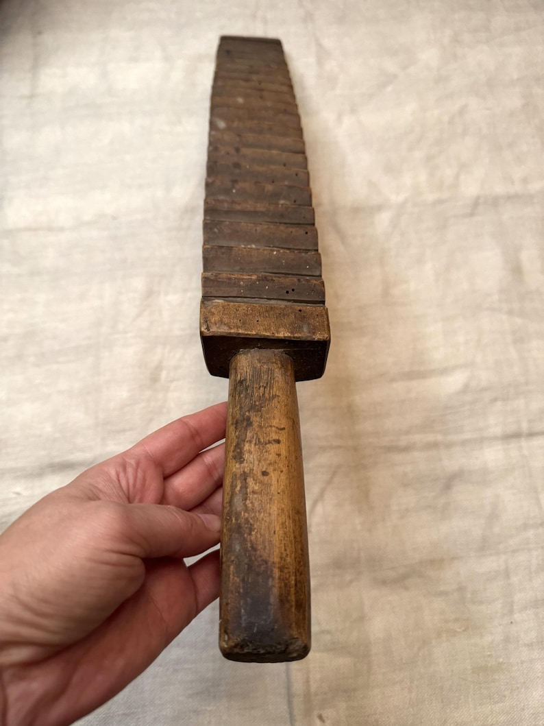 Pu&ograve; includere: Un lungo attrezzo di legno con un manico e una serie di blocchi rettangolari impilati. L'attrezzo &egrave; in legno marrone scuro e ha un aspetto usurato e rustico.