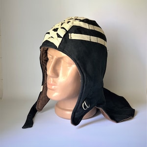 Puede incluir: Sombrero de lana negro con un diseño tejido blanco en la parte superior y un forro de lana marrón. El sombrero tiene una solapa larga que se puede atar debajo de la barbilla.