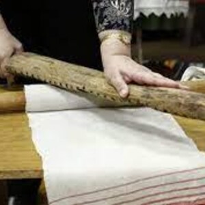 Pu&ograve; includere: Una donna usa un matterello di legno con un bordo seghettato per lisciare un panno di lino bianco su un tavolo di legno.