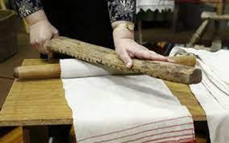 Pu&ograve; includere: Una donna usa un matterello di legno con un bordo seghettato per lisciare un panno di lino bianco su un tavolo di legno.