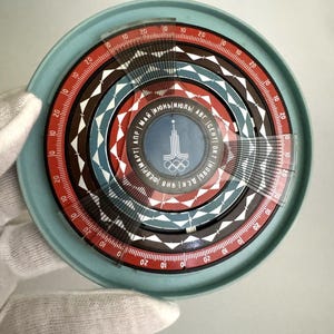 Puede incluir: Un calendario circular de plástico azul con un diseño rojo, blanco y negro. El calendario presenta los anillos olímpicos y el texto "НОЯБ ДЕК ЯНВ ФЕВРМАРТ АПР МАЙ ИЮНЬ ИЮЛЬ АВГ СЕНТ ОКТ".