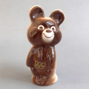 Könnte beinhalten: Eine glänzende braune Keramikbärenfigur mit weißem Gesicht, Ohren und Brustbereich. Der Bär hat einen lächelnden Ausdruck und zeigt das goldene Symbol der olympischen Ringe auf seinem Bauch. Die Figur steht aufrecht mit einer glatten Oberfläche.