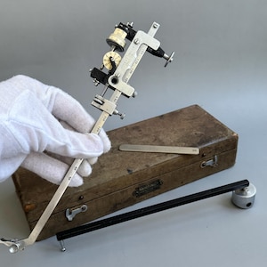 May include: A vintage silver and black metal planimeter instrument featuring a graduated scale, rotating dials, and adjustment screws. The device is accompanied by a rectangular wooden storage case with metal latches and a separate black metal arm.