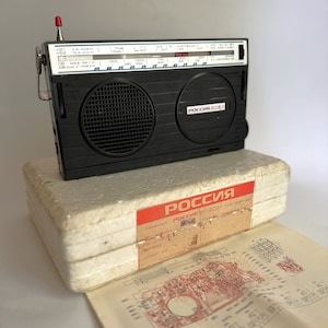 May include: A black and silver vintage radio with a white dial and the text "РОССИЯ 203-1" on the front. The radio is in its original white foam packaging with a red label that says "РОССИЯ РП-203-1 гост 3-720-81".