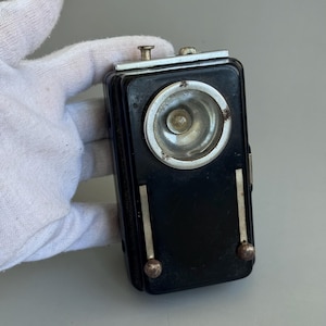 May include: A vintage black flashlight with a circular silver bezel around the light. The flashlight has a rectangular shape with silver accents and two small, round, silver-colored knobs at the bottom. The flashlight is held by a gloved hand.