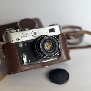 May include: A silver and black 35mm film camera with a brown leather case. The camera has a lens with the text "FED 3" and "28/3.5" printed on it.