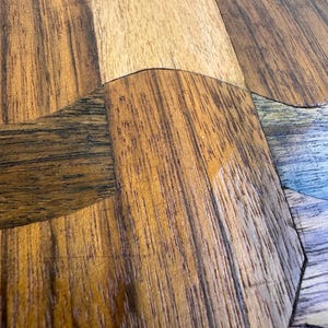 Peut inclure: Gros plan sur une surface en bois avec un motif g&eacute;om&eacute;trique. Le bois pr&eacute;sente une vari&eacute;t&eacute; de tons chauds, dont le beige clair, le marron moyen et le marron fonc&eacute;, dispos&eacute;s dans un motif en mosa&iuml;que. Le grain du bois est visible.