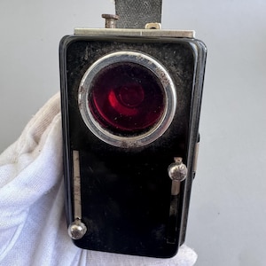 May include: A vintage black lantern with a red lens and silver trim. The lantern has a rectangular shape with a handle and a small metal clasp. The lens is circular and appears to be made of glass. The lantern is likely from the early 20th century.