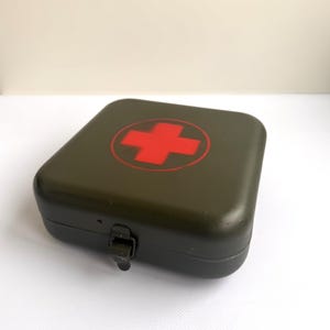Puede incluir: Un botiquín verde oliva con una cruz roja en la tapa. La caja cuadrada tiene un cierre de metal. Este kit está diseñado para guardar suministros médicos y es adecuado para el hogar o viajes.