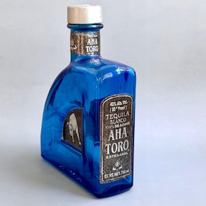 Puede incluir: Una botella de tequila de vidrio azul cobalto con tapa plateada. La etiqueta dice AHA TORO TEQUILA BLANCO 100% DE AGAVE, 40% Alc. Vol. (80 Proof) y CONT. NET. 750 ml. Un gráfico de un toro plateado está grabado en el costado de la botella.