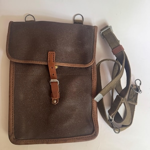 May include: A brown leather satchel with a flap closure and a detachable olive green strap. The satchel has a rectangular shape with a lighter brown trim and a buckle closure. Two metal rings are attached to the top of the satchel.