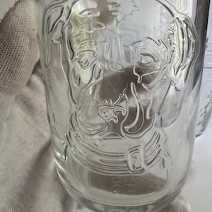 May include: Clear glass tumbler with an embossed image of a dog's face. The dog's features are detailed, including its eyes, nose, and ears. The glass is smooth and transparent, allowing the contents to be visible. The tumbler is a standard size for beverages.