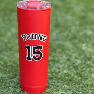 May include: A matte red cylindrical tumbler with a clear plastic lid. The front of the tumbler features the name YOUNG in black letters with a white outline, positioned above the number 15 in a matching black and white athletic font style.