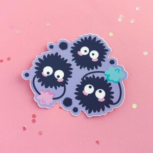 May include: A sticker featuring three black and white fuzzy creatures with large eyes and pink cheeks. The creatures are clustered together on a purple background with a green leaf in the upper right corner.