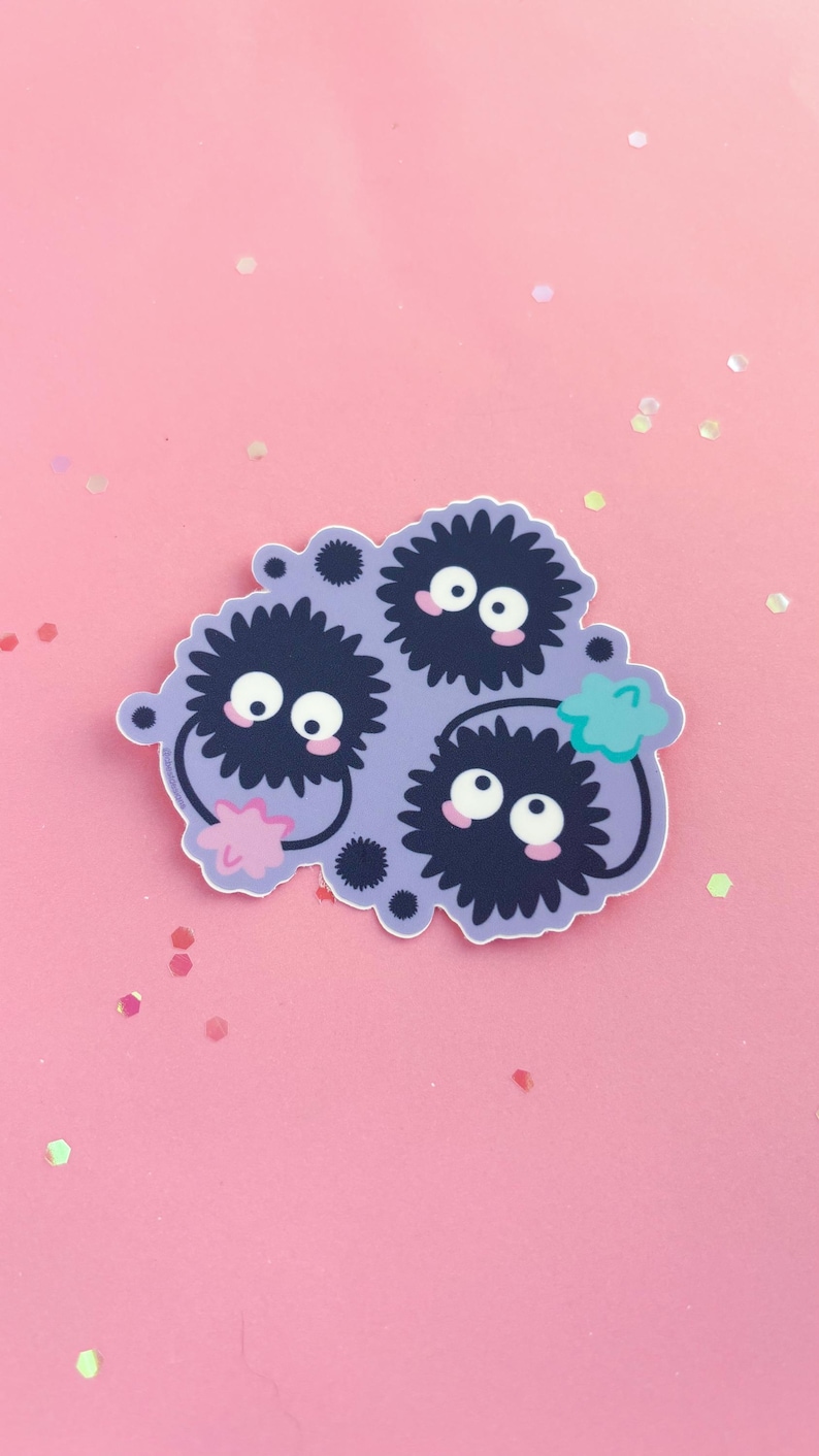 May include: A sticker featuring three black and white fuzzy creatures with large eyes and pink cheeks. The creatures are clustered together on a purple background with a green leaf in the upper right corner.