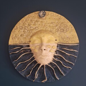Może przedstawiać: Ceramiczna dekoracja ścienna w kształcie koła. Górna połowa jest teksturowana na żółto, dolna połowa na niebiesko z promienistymi liniami. Stylizowana twarz z zamkniętymi oczami znajduje się pośrodku. Mały, ciemny, okrągły akcent znajduje się na górze.