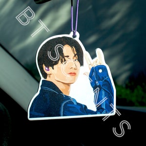May include: A white car air freshener with a cartoon illustration of a man with black hair wearing a blue denim jacket. The man is making a peace sign with his right hand. The air freshener is hanging from a rearview mirror.