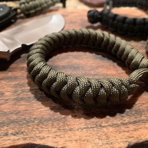 Puede incluir: Un brazalete de paracord verde de cerca sobre una superficie de madera. El brazalete está hecho de paracord trenzado y tiene un nudo simple en el extremo.
