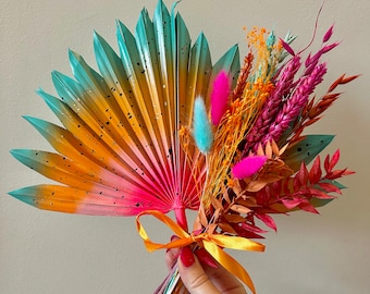 May include: A colorful bouquet of dried flowers and grasses, featuring a large, painted palm leaf fan. The bouquet is tied with a yellow ribbon and includes pink, orange, and blue dried flowers.