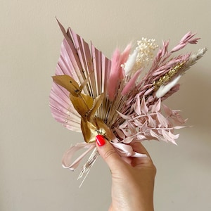 May include: A bouquet of dried flowers in shades of pink and white, with gold accents. The bouquet includes palm leaves, pampas grass, and other dried flowers.