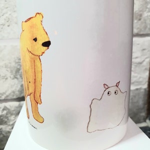 Könnte beinhalten: Eine weiße Keramiktasse mit einer Illustration eines gelben Bären und eines weißen Geistes. Der Bär steht auf seinen Hinterbeinen und der Geist schwebt in der Luft. Die Tasse hat eine schwarze Signatur am Boden.