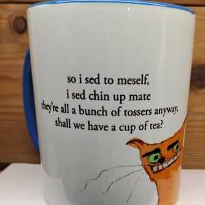 Puede incluir: Taza de cerámica blanca con asa y borde azules. La taza presenta una ilustración de gato de dibujos animados y el texto: "so i sed to meself, i sed chin up mate they're all a bunch of tossers anyway, shall we have a cup of tea?"