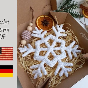 May include: A white crocheted snowflake ornament rests in a cardboard box filled with straw. The snowflake is the focal point, with intricate details. The box also contains dried orange slices and wooden beads. Text on the image reads "Crochet pattern PDF".