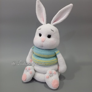 Può includere: Coniglio bianco all'uncinetto con orecchie lunghe, che indossa un maglione a righe blu e gialle. Il coniglio è seduto con le zampe ripiegate sotto il corpo. Il coniglio è un giocattolo amigurumi fatto a mano.