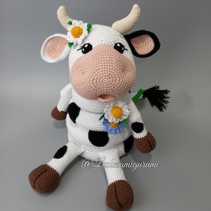 Könnte beinhalten: Ein gehäkeltes Stofftier-Kuh mit schwarzen und weißen Flecken, braunen Hufen und einer rosa Nase. Die Kuh trägt eine weiße Blumenkrone und eine Blumenkette.