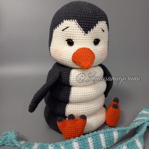 Könnte beinhalten: Ein gehäkeltes Pinguin-Spielzeug mit weißem Körper, schwarzen Flügeln und einem orangefarbenen Schnabel. Der Pinguin trägt einen blau-weiß gestreiften Schal. Das Spielzeug sitzt auf einer grauen Oberfläche. @Ludasamigurumi