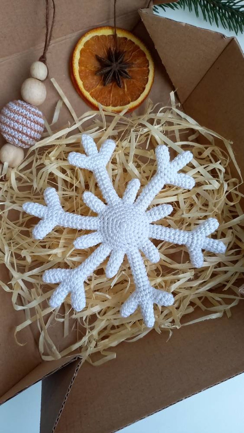 May include: A white crocheted snowflake ornament in a box with shredded paper. The snowflake is a popular Christmas decoration.