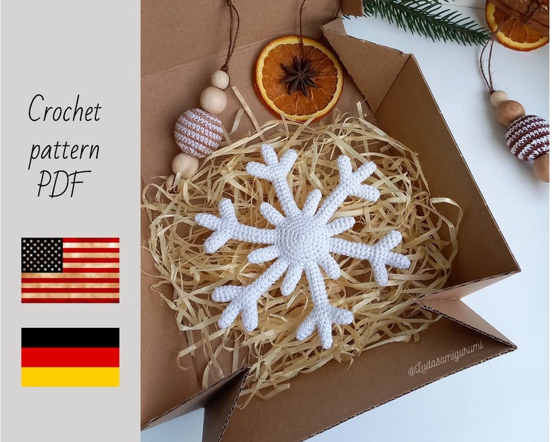 May include: A white crocheted snowflake ornament rests in a cardboard box filled with straw. The box also contains dried orange slices and wooden beads. The text "Crochet pattern PDF" is visible, along with the American and German flags.