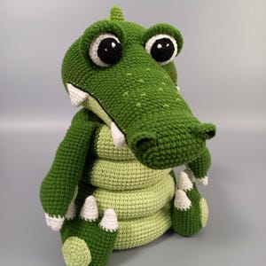 Op de afbeelding: Een handgemaakt gehaakt alligatorspeelgoed in groen en lichtgroen, met grote zwart-witte ogen en witte tanden. De alligator heeft een getextureerd, gestikt uiterlijk en zit rechtop. Het speelgoed is een grillig, handgemaakt item.