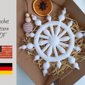 May include: A white crochet snowflake ornament with a detailed design, displayed in a cardboard box with straw. The image includes text that reads "Crochet pattern PDF" and flags of the USA and Germany.