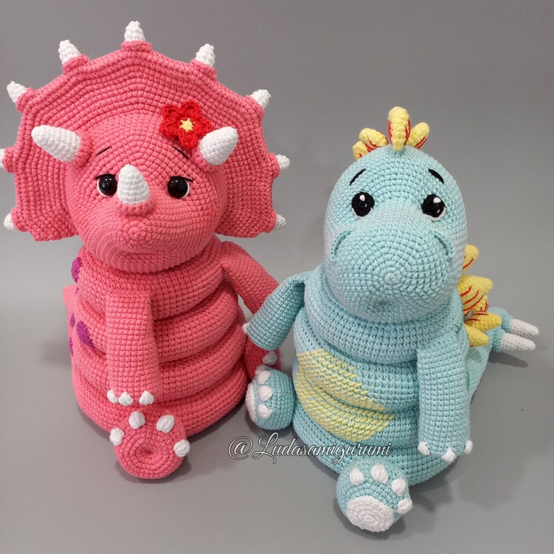 May include: Two crocheted dinosaur toys, one pink with a flower on its head and one light blue with a yellow heart on its chest. The toys are sitting on a gray surface.