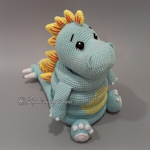 May include: A crocheted dinosaur toy with a light blue body, yellow and red spikes, and white feet. The toy has a cute, cartoon-like face with black eyes and a small mouth.