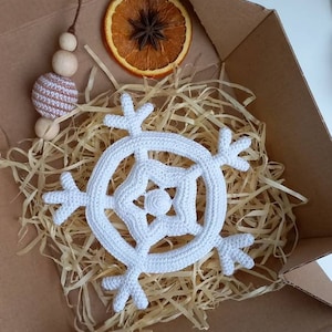 May include: A white crocheted snowflake ornament in a cardboard box with brown packing paper. The ornament is a simple design with six points and a small circle in the center.