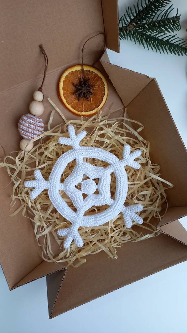May include: A white crocheted snowflake ornament in a cardboard box with brown packing paper. The ornament is a simple design with six points and a small circle in the center.