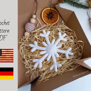 May include: A white crocheted snowflake ornament in a brown cardboard box with shredded paper. The box is open and the snowflake is visible. The text "@lyudasamigurumi" is visible on the bottom right corner of the box.