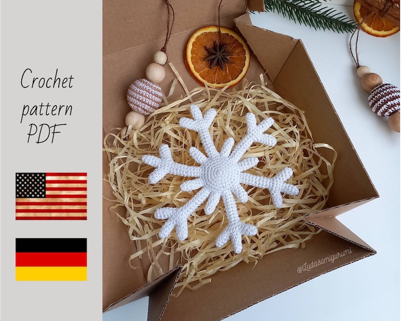 May include: A white crocheted snowflake ornament in a brown cardboard box with shredded paper. The box is open and the snowflake is visible. The text "@lyudasamigurumi" is visible on the bottom right corner of the box.