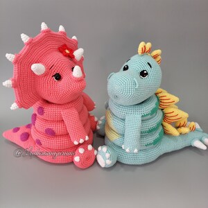 May include: Two crocheted dinosaur toys, one pink with purple spots and a flower on its head, and one blue with yellow stripes. The toys are sitting on a light gray surface.