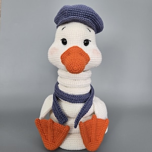 Op de afbeelding: Een handgemaakte gehaakte ganzenpop met een witte body, oranje snavel en voeten, en een blauwe hoed en sjaal. De gans heeft zwarte ogen en roze wangen, wat een grillig en charmant uiterlijk creëert. Een uniek, handgemaakt item.