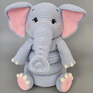 Pode incluir: Um elefante de pelúcia feito em crochê na cor azul claro com orelhas grandes que possuem painéis internos cor-de-rosa. O elefante tem olhos de segurança pretos, uma tromba longa e quatro patas com detalhes brancos nos dedos e almofadas rosas.